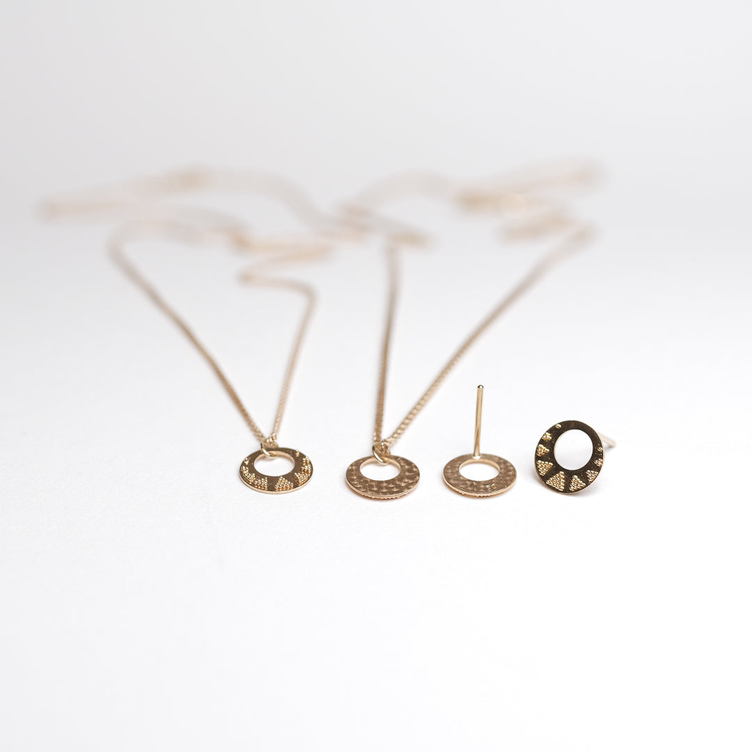 Das Bild zeigt zwei Kettenanhänger welche durch Granulationstechnik verziert sind. Die Anhänger hängen an jeweils einer feinen Zopfkette und werden vor einem Stück Baumrinde präsentiert. Zusätzlich liegt noch ein Paar granulierter Ohrstecker auf dem Papieruntergrund.