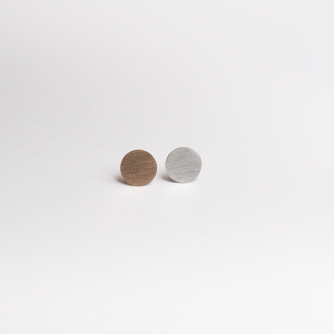 Das Bild zeigt runde und flache Ohrstecker. der linke ist aus Gold, der rechte ist aus Platin. Beide Ohrstecker liegen auf einer weißen Papieroberfläche