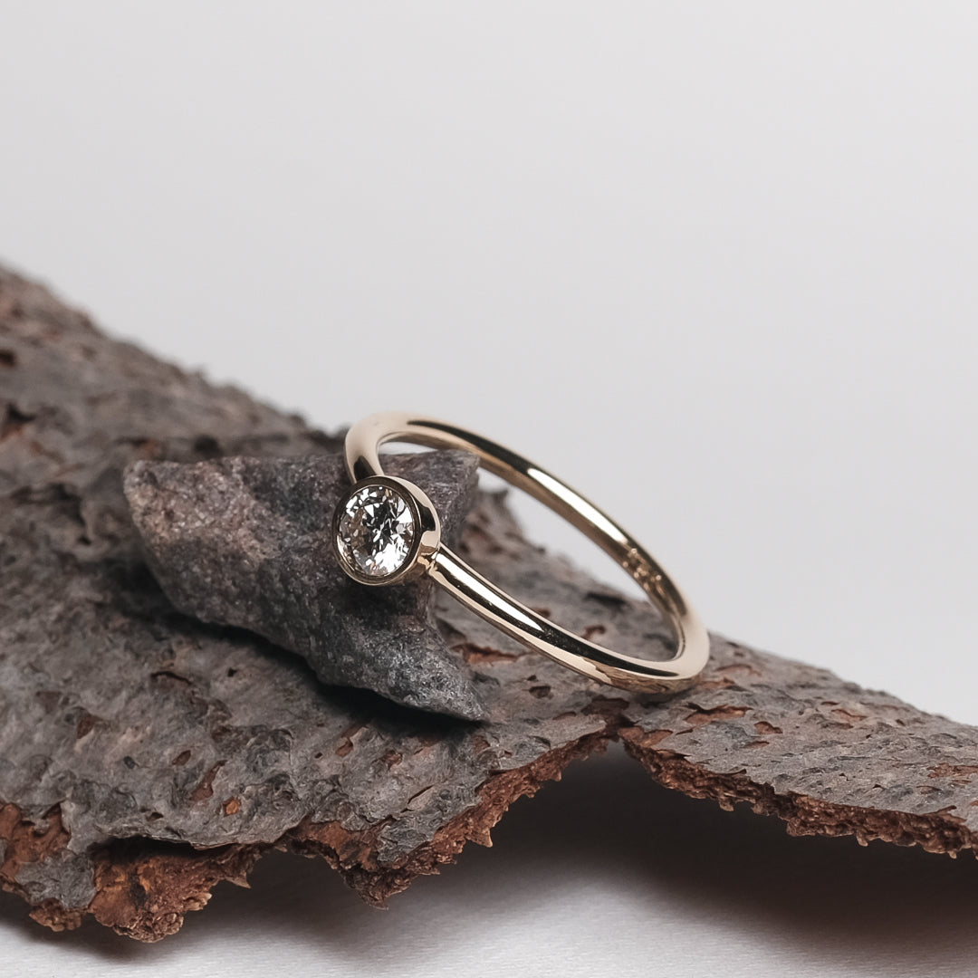 Das Bild zeigt den Brillant Ring aus der Produktbeschreibung. Es zeigt einen Gold Ring weicher mit einem Brillant besetzt ist. Der Ring wird auf einer Holz Unterlage präsentiert.