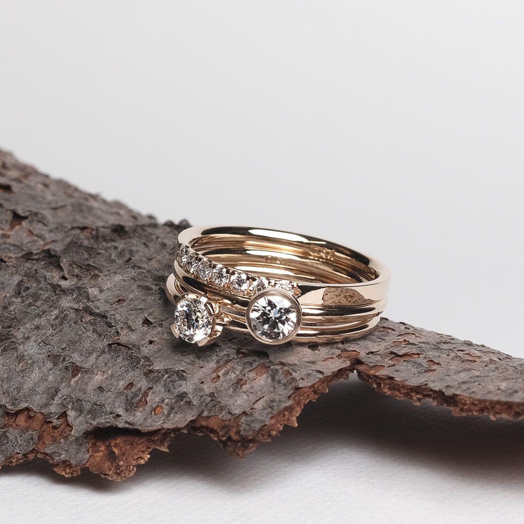 Das Bild zeit eine Sammlung aus Brillantringen aus Gelbgold. Der oberste ist der Ring mit sieben in Reihe gefassten Brillanten. Der Ring ist auf einem Stück Baumrinde platziert, welche auf einem hellen Papieruntergrund präsentiert wird.