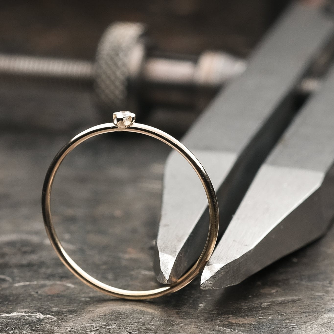 Sehr feiner Brillantring, welcher auf einem dunklen Amboss präsentiert wird. Der Ring klemmt in einem Metall Zirkel, welcher auf dem Amboss liegt. Zu erkennen ist der sehr filigrane Brillant des Rings, welcher in einer Vierkrappenfassung gefasst ist.