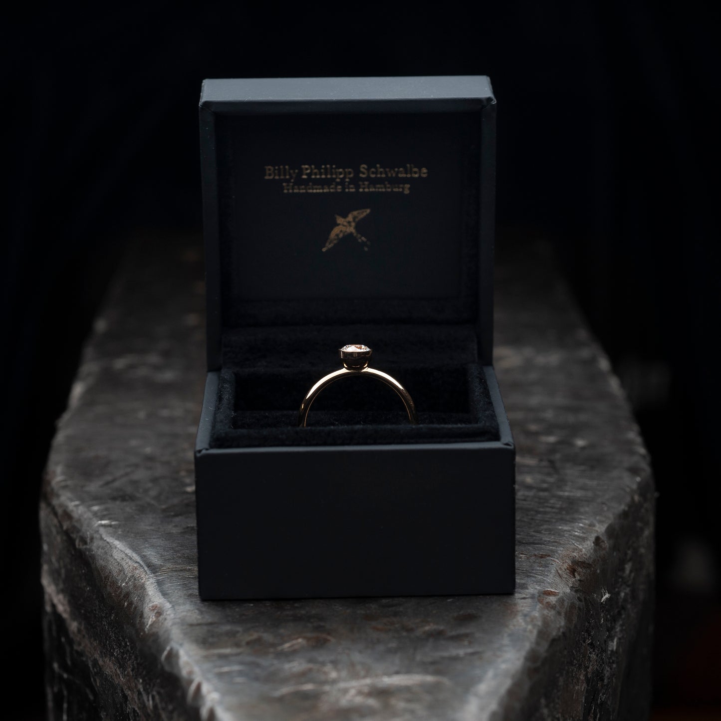 Der Ring liegend auf einem massiven Amboss. Ring steckt in einer schwarzen Schmuckschachtel. 