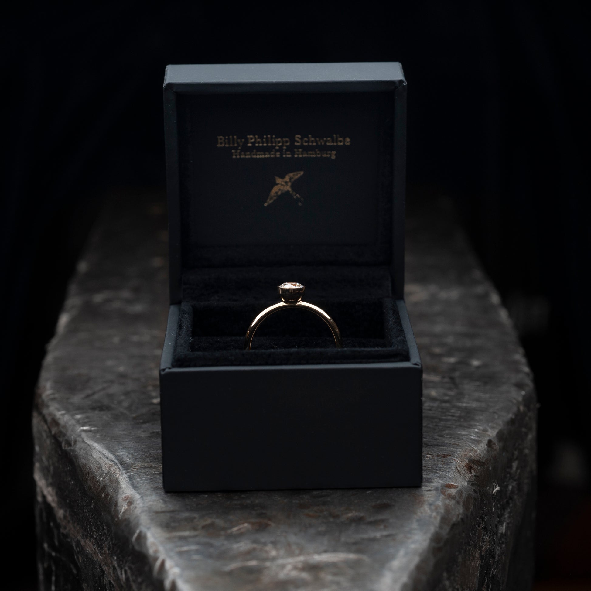 Der Ring liegend auf einem massiven Amboss. Ring steckt in einer schwarzen Schmuckschachtel. 