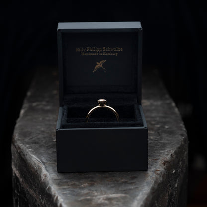 Der Ring liegend auf einem massiven Amboss. Ring steckt in einer schwarzen Schmuckschachtel. 
