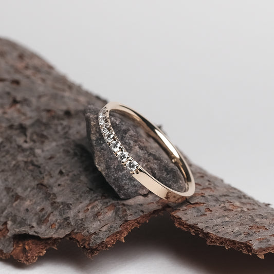 Das Bild zeit einen Brillantring aus Gelbgold, mit sieben in Reihe gefassten Brillanten. Der Ring ist auf einem Stück Baumrinde platziert, welche auf einem hellen Papieruntergrund präsentiert wird.