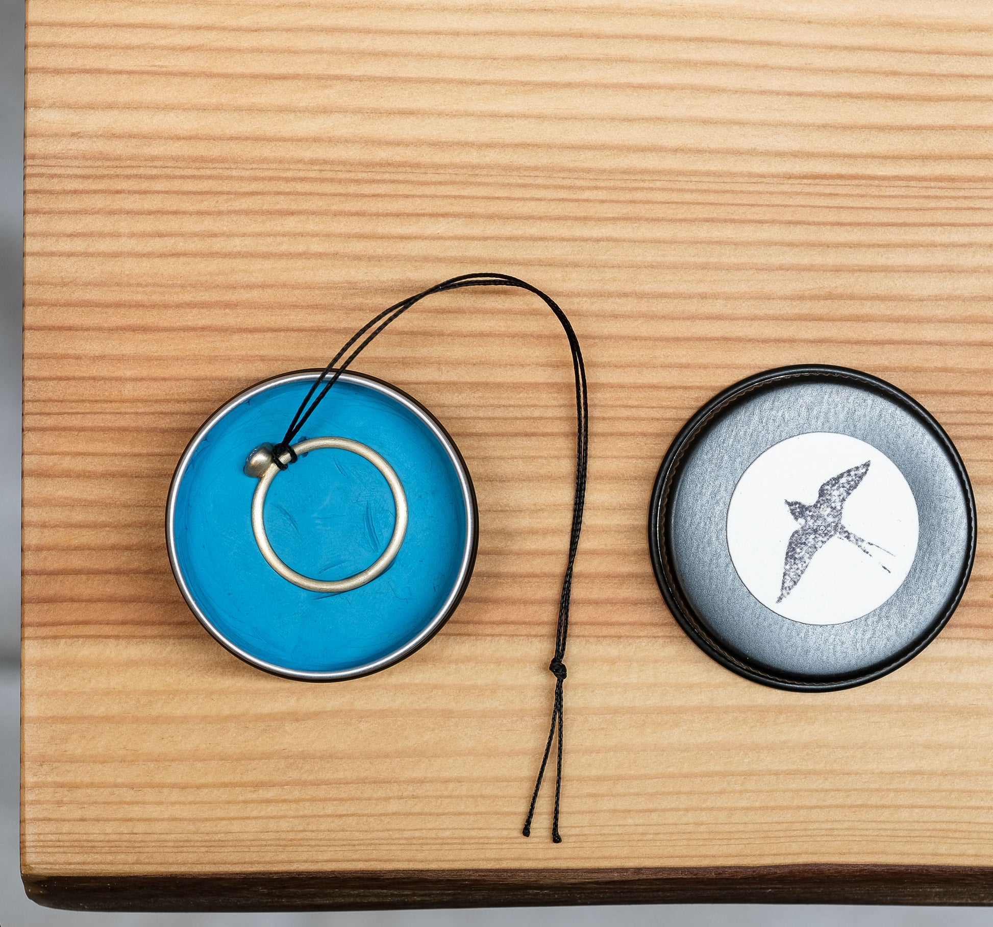 Ringgrößen-Spionageset bestehend aus einer Dose mit der Abdruckmasse und mit schwarzem Band zum wieder herausziehen des Eingedrückten Rings. Die runde Dose enthält die blaue Abdruckmasse. Daneben der Deckel mit einem Schwalbenmotiv auf hellem Etikett, auf einer Holzoberfläche liegend.