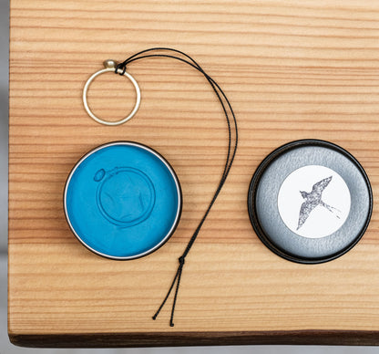 Ringgrößen-Spionageset bestehend aus einer Dose mit der Abdruckmasse und mit schwarzem Band zum wieder herausziehen des Eingedrückten Rings. Die runde Dose enthält die blaue Abdruckmasse. Daneben der Deckel mit einem Schwalbenmotiv auf hellem Etikett, auf einer Holzoberfläche liegend.