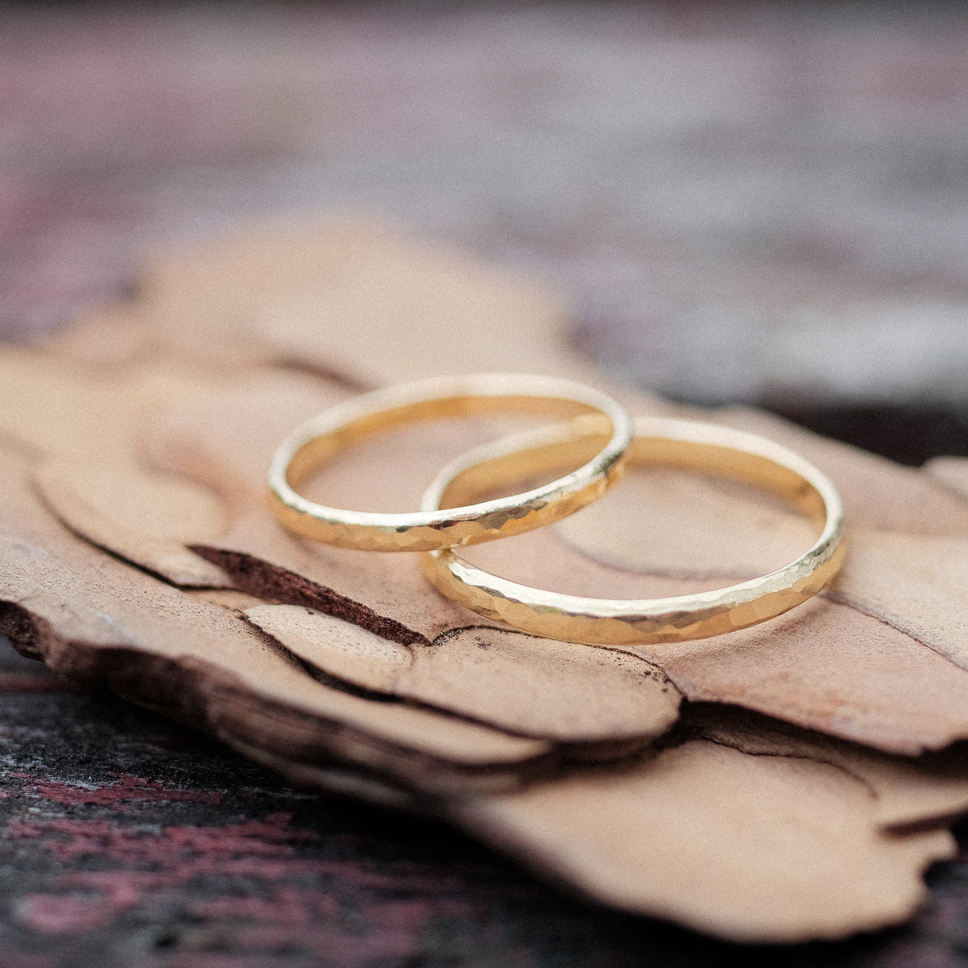 Das Bild zeigt feine Gold Eheringe welche außen halbrund und gehämmert sind und innen Flach und poliert sind. Sie werden auf einer Holzoberfläche liegend präsentiert. 