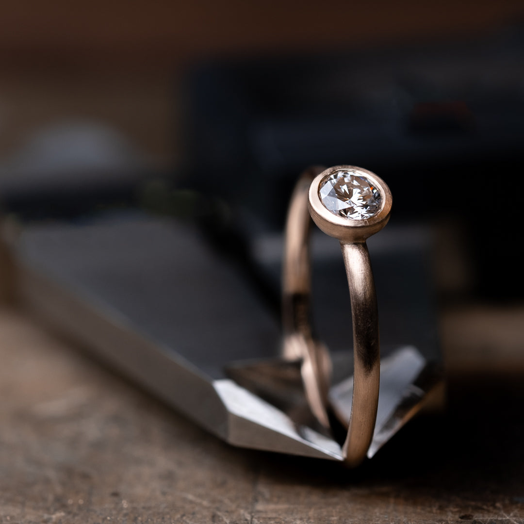 Das Foto Zeit einen Brillantring aus Rose Gold, welcher einen 0,25ct. großen Brillanten in Zargenfassung beeinhaltet.
Der Ring ist mattiert und wird eingeklemmt in einem Messschieber präsentiert.