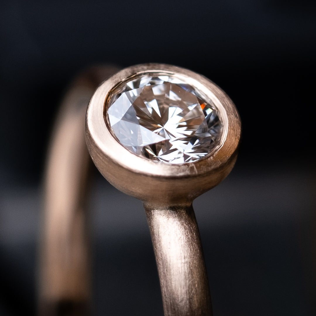 Das Foto Zeit einen Brillantring aus Rose Gold, welcher einen 0,25ct. großen Brillanten in Zargenfassung beeinhaltet.
Der Ring ist mattiert. Es handelt sich um eine Macro Fotografie des eingesetzten Diamanten und dessen Fassung.