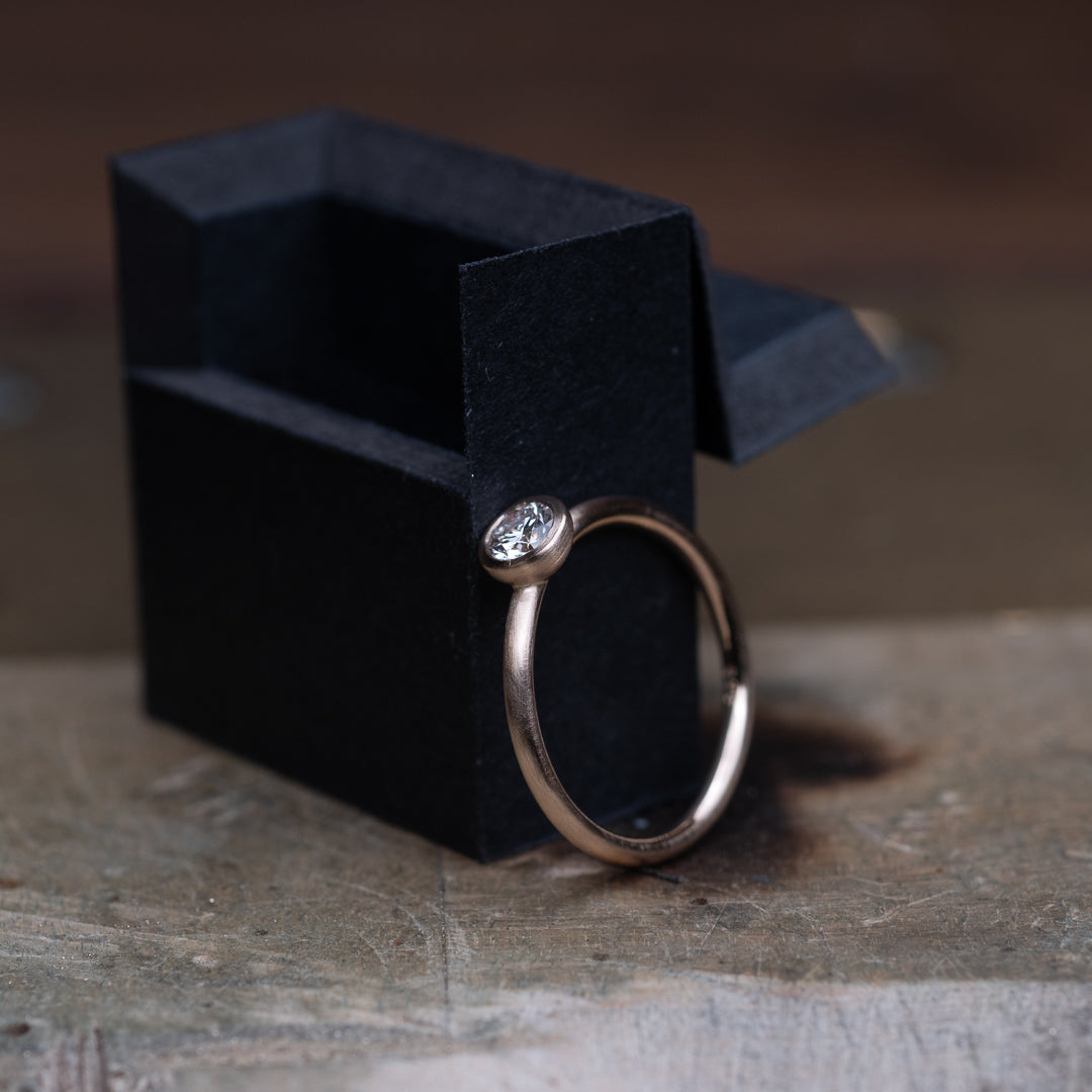 Das Foto Zeit einen Brillantring aus Rose Gold, welcher einen 0,25ct. großen Brillanten in Zargenfassung beeinhaltet.
Der Ring ist mattiert. Der Ring lehnt an einer kleinen Ringschachtel auf einer Fäche aus Holz.