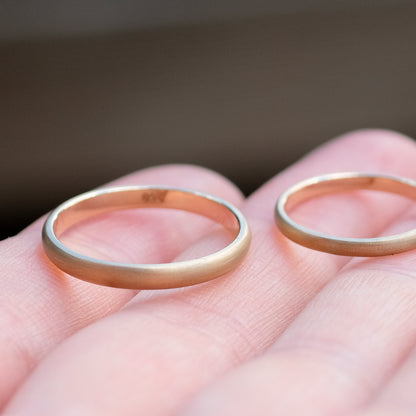 Das Bild zeigt feine Gold Eheringe welche außen halbrund und mattiert sind und innen Flach und poliert sind. Sie werden auf einer Handoberfläche liegend präsentiert. 