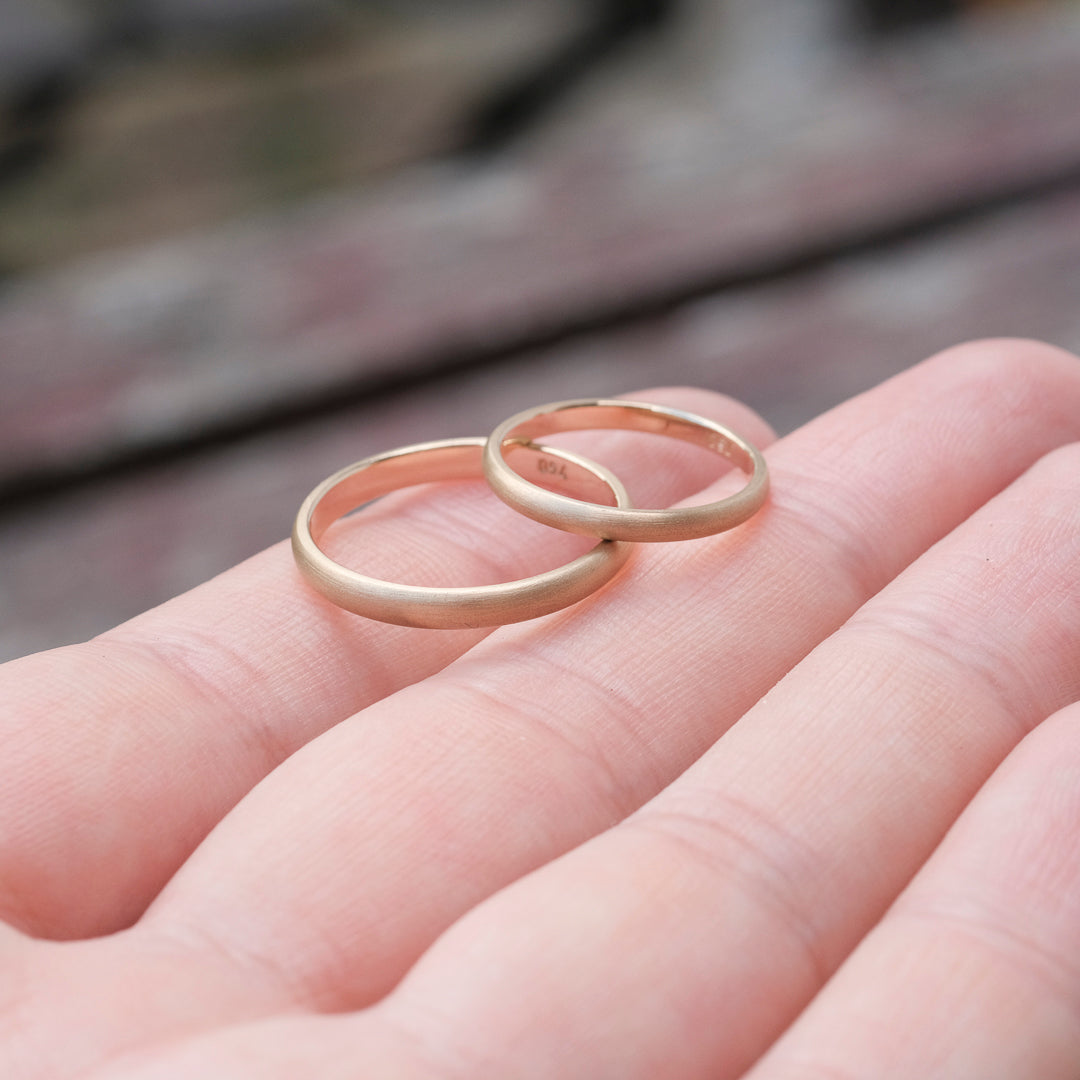 Das Bild zeigt feine Gold Eheringe welche außen halbrund und mattiert sind und innen Flach und poliert sind. Sie werden auf einer Handoberfläche liegend präsentiert. 