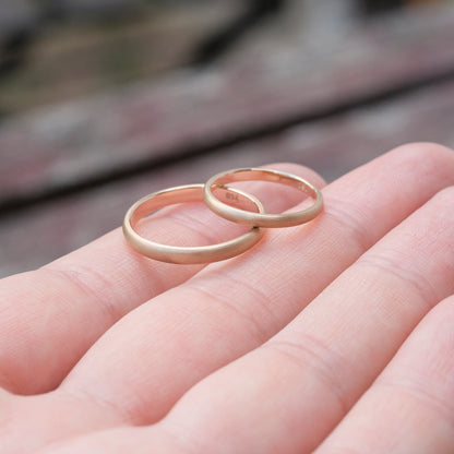 Das Bild zeigt feine Gold Eheringe welche außen halbrund und mattiert sind und innen Flach und poliert sind. Sie werden auf einer Handoberfläche liegend präsentiert. 