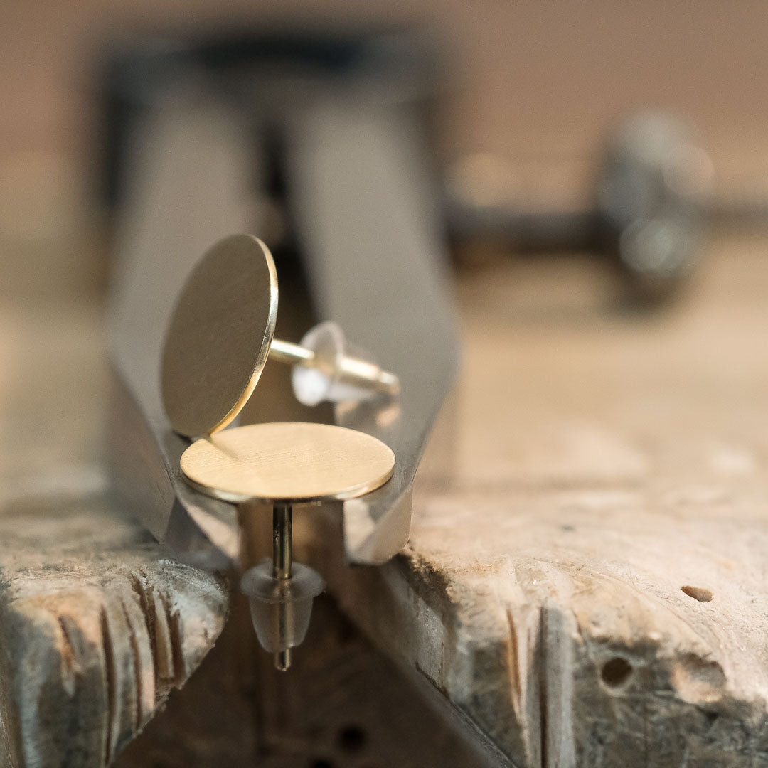Das Bild zeigt runde und flache Ohrstecker aus Gold, welche auf einem Stahl Zirkel liegend präsentiert werden. 