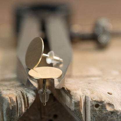 Das Bild zeigt runde und flache Ohrstecker aus Gold, welche auf einem Stahl Zirkel liegend präsentiert werden. 